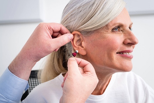 Ältere Frau bekommt ein modernes Hörgerät ins Ohr eingesetzt, Hörakustiker hilft dabei.